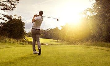 A golfer swings a club on a grassy course, facing lush greenery and a bright, setting sun.