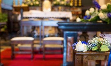 Two wooden chairs facing an altar adorned with flowers and candles, within a brightly lit, decorated church interior, featuring a red carpet and floral arrangements on the side.