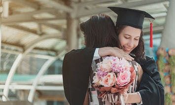 A graduate in a cap and gown hugs another person while holding a bouquet of flowers, standing under a glass-covered structure.