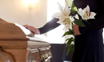 A person in black attire holds white lilies and rests a hand on a wooden casket in a softly lit room, conveying a somber and respectful mood.