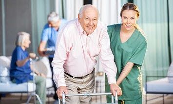 An elderly man using a walker receives assistance from a smiling caregiver in a green uniform. In the background, two other elderly people sit and engage with their own caregivers.