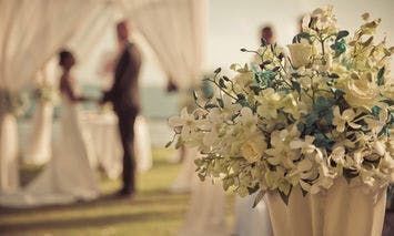 Bouquet of white flowers in a vase with a couple standing and holding hands in the center background, framed by white drapery, during an outdoor event.
